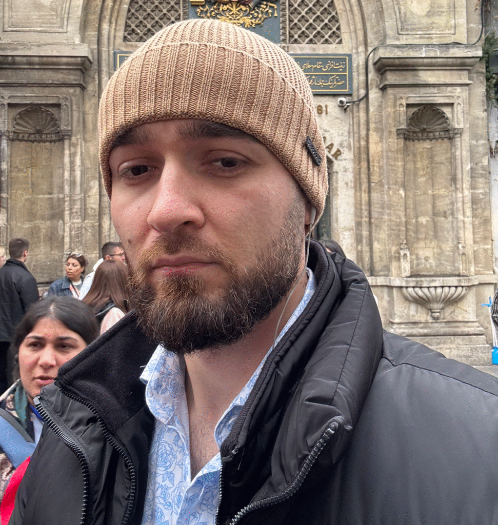 A man with a trimmed beard and mustache wearing a tan knit beanie and a black jacket over a patterned shirt, standing outdoors in front of an ornate stone building with people in the background.